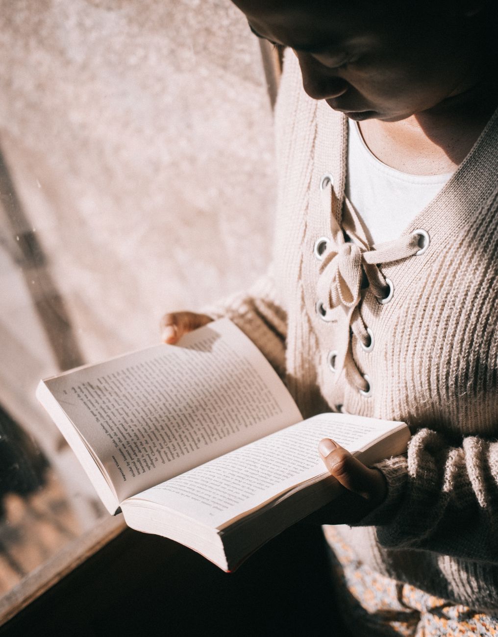 A person reading a book while sitting by a window, wearing a cozy knitted sweater.